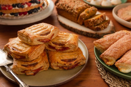 A variety of baked goods, including flaky pastries, a layered cake, and slices of bread, arranged on wooden table.