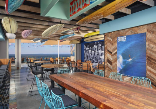 A modern restaurant interior with wooden tables, colorful surfboards hanging, and ocean views.