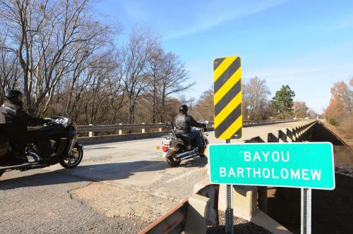 Two motorcyclists ride past a sign for Bayou Bartholomew on a bridge surrounded by bare trees.