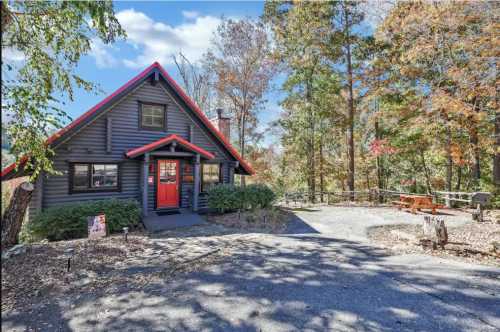 A charming log cabin with a red roof and door, surrounded by autumn trees and a gravel driveway.