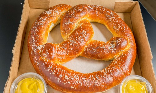 A large, golden-brown pretzel with coarse salt, served in a box with two cups of mustard on the side.