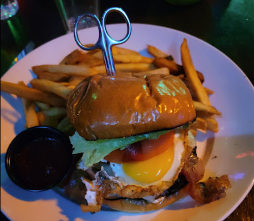 A burger with a fried egg, bacon, lettuce, and tomato, served with fries and a side of dipping sauce.