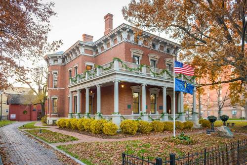 Historic brick mansion adorned with greenery, surrounded by autumn foliage and flags, set in a peaceful neighborhood.