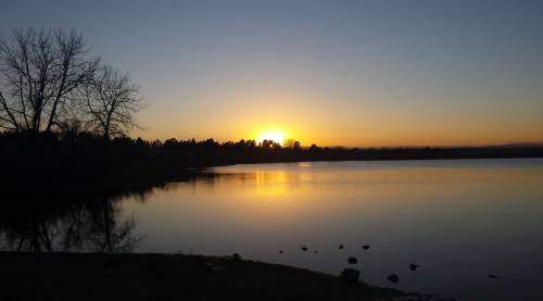 A serene sunset over a calm lake, with silhouettes of trees and a clear sky reflecting on the water's surface.