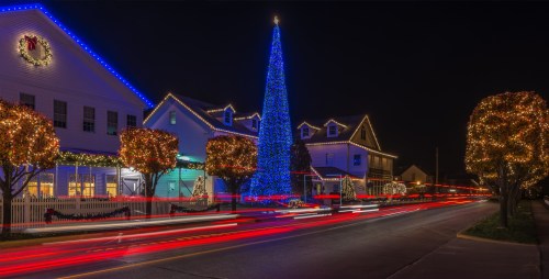 A brightly lit Christmas tree stands in a festive town square, surrounded by decorated buildings and light trails from passing cars.