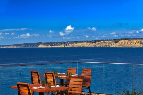 A seaside restaurant patio with wooden tables overlooking a calm blue ocean and distant cliffs under a clear sky.