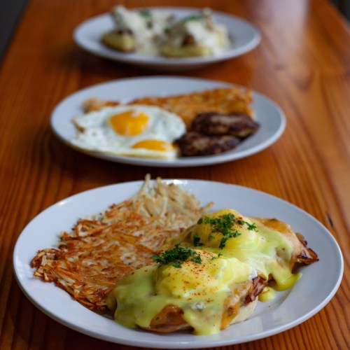 Three plates of breakfast: eggs Benedict with hash browns, sunny-side-up eggs with sausage, and a potato dish in the background.