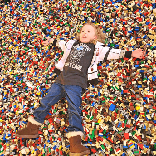 A child lies on a colorful pile of LEGO bricks, arms outstretched, smiling happily.