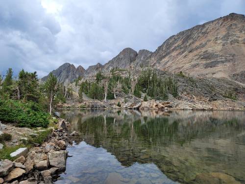 A serene mountain lake reflecting rocky peaks and lush greenery under a cloudy sky.