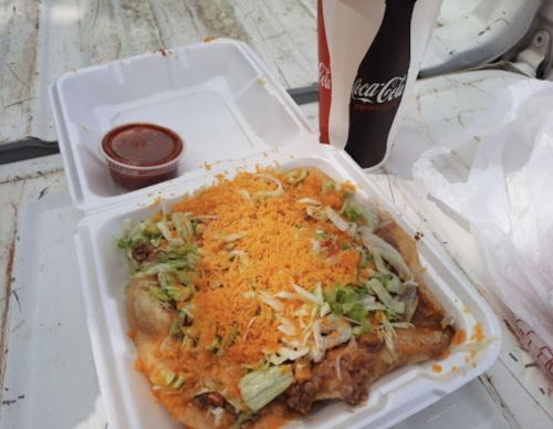 A takeout container with a loaded taco topped with lettuce, cheese, and a side of salsa, next to a Coca-Cola.