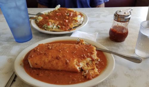 A plate with a smothered burrito in sauce, alongside a plate of hash browns topped with vegetables and a bottle of hot sauce.