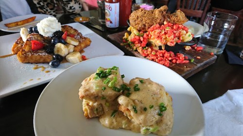 A plate of French toast topped with fruit, a dish of fried chicken with colorful toppings, and biscuits with gravy.