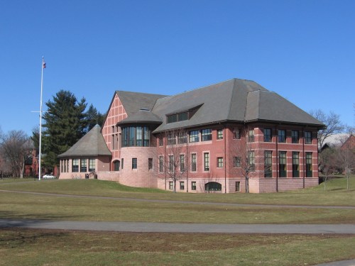 A large, red-brick building with a sloped roof, surrounded by green grass and trees under a clear blue sky.