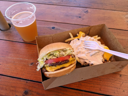 A burger with lettuce and tomato, a side of cheesy fries, and a cup of beer on a wooden table.