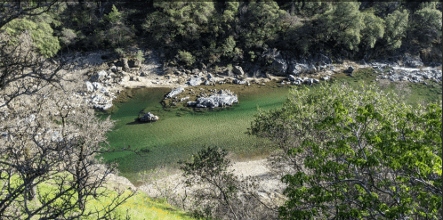 A serene riverbank scene with clear green water, rocky formations, and lush greenery surrounding the area.