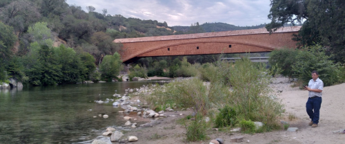 A covered bridge spans a river, with a man walking along the shore and lush greenery surrounding the scene.