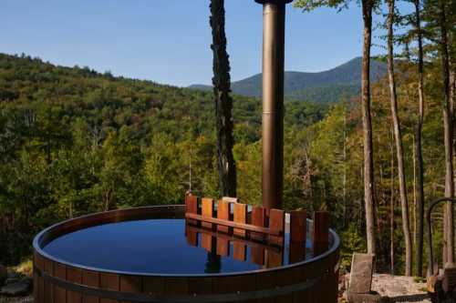 A wooden hot tub sits outdoors, surrounded by lush green trees and mountains under a clear blue sky.