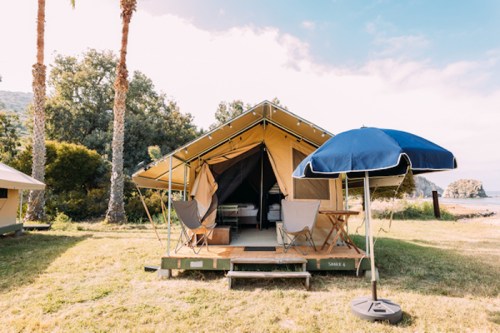 A glamping tent with a porch, chairs, and an umbrella, set in a grassy area with trees and a scenic backdrop.