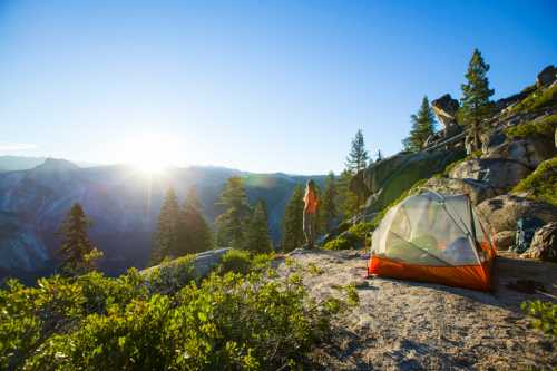 A person stands near a tent on a rocky hillside, overlooking a valley at sunrise, surrounded by trees.