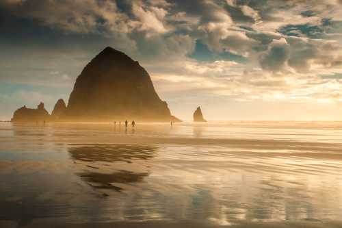 Silhouetted figures walk along a sandy beach at sunset, with dramatic rock formations and clouds in the background.