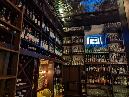 A cozy bar with wooden shelves filled with various bottles of liquor and glassware, illuminated by soft lighting.