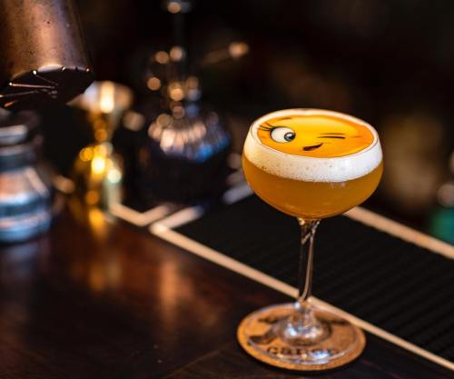 A cocktail in a coupe glass with a playful emoji design on the foam, set on a bar counter.