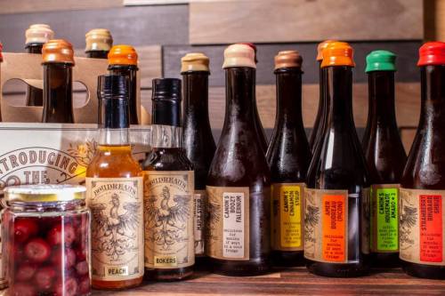 A variety of craft beer bottles with colorful caps, alongside a jar of cranberries and a bottle of peach syrup.