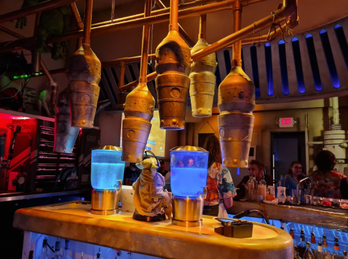 A futuristic bar with glowing blue drinks and metallic decor, featuring unique drink dispensers and patrons in the background.