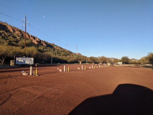 A dirt road lined with yellow posts, surrounded by mountains and a clear blue sky with a visible moon.