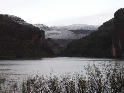 A serene lake surrounded by rocky cliffs and snow-capped mountains under a cloudy sky.