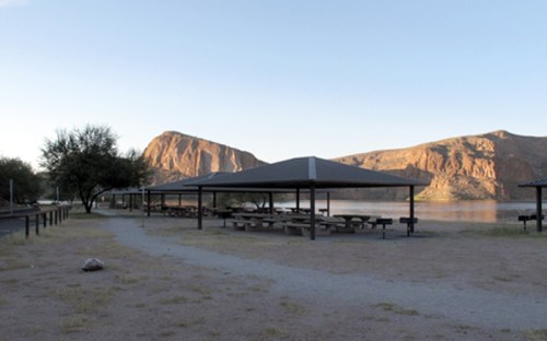 A serene lakeside park with picnic shelters and mountains in the background under a clear sky.