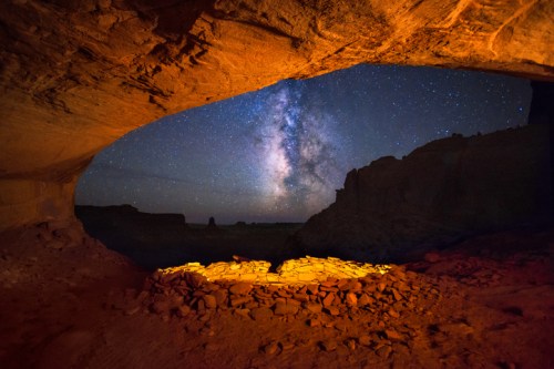 A cave opening reveals a starry night sky with the Milky Way, illuminated by warm orange light on the rocky ground.