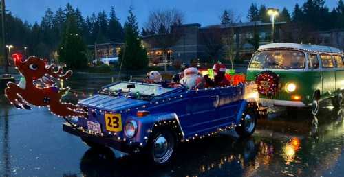 A decorated blue convertible with Santa and reindeer, alongside a green vintage van, in a festive, rainy setting.