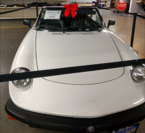 A white convertible car with a red bow on top, displayed behind a barrier in a showroom. Price tag visible.