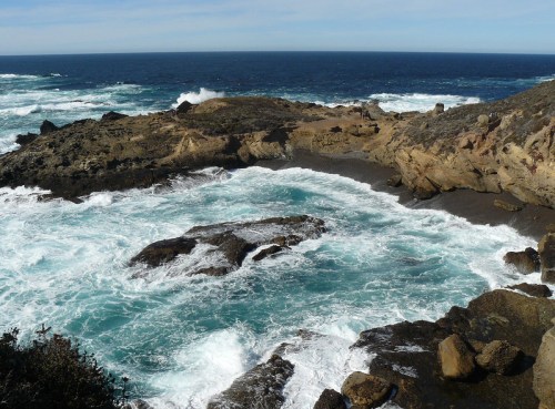 A rocky coastline with waves crashing against the shore, creating a turquoise lagoon under a clear blue sky.