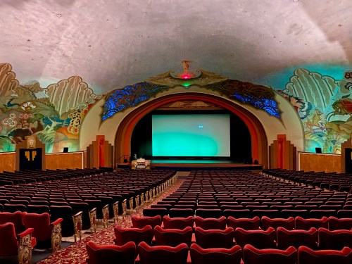 A vintage theater interior with red seats, a large screen, and colorful murals on the ceiling and walls.