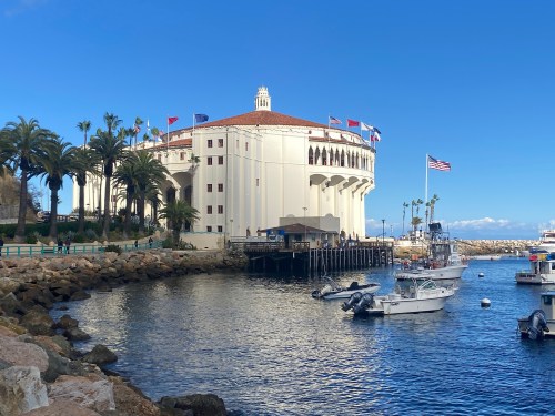 A scenic view of a waterfront building with flags, palm trees, and boats in a calm harbor under a clear blue sky.