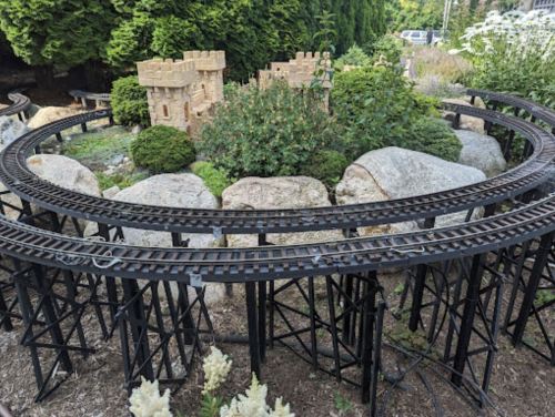 A miniature train track curves around rocks and greenery, featuring a small castle structure in the background.