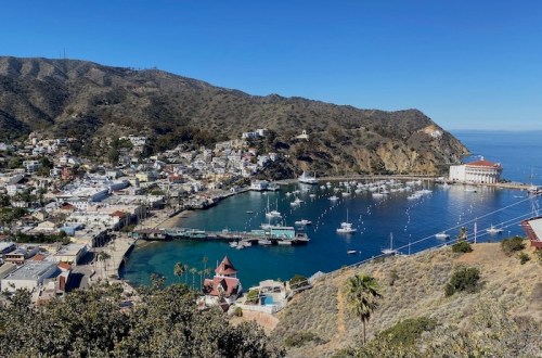A scenic view of a coastal town with boats in the harbor, surrounded by hills and clear blue skies.