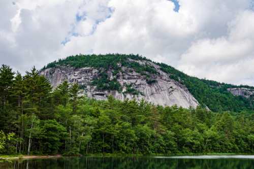 A rocky mountain rises above a lush green forest, reflecting in a calm lake under a cloudy sky.