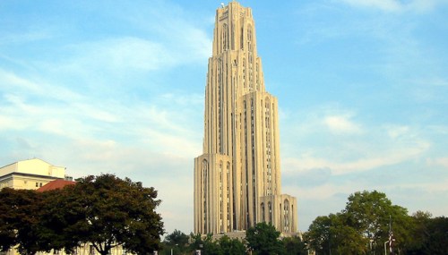 A tall, ornate building with a spire, surrounded by trees and a blue sky.