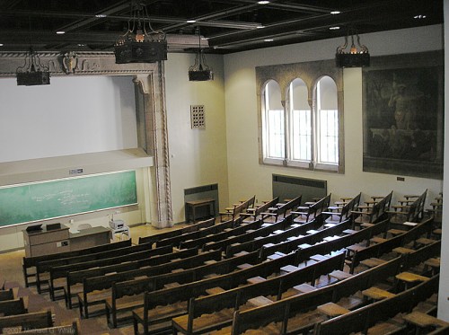 A spacious lecture hall with wooden seating, a chalkboard, and large windows, featuring a decorative ceiling and wall art.
