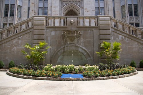 A decorative fountain surrounded by colorful flowers and plants, set against a grand stone building.