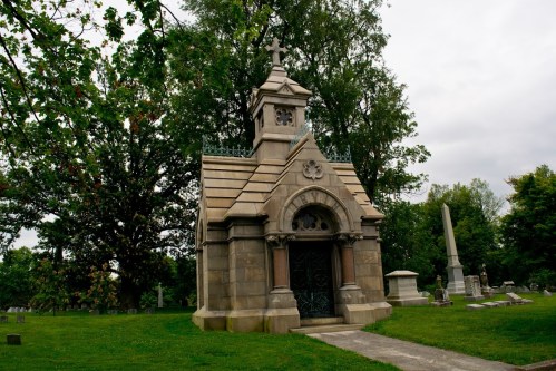A stone mausoleum with a cross on top, surrounded by trees and gravestones in a cemetery. Overcast sky above.