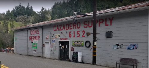 A gray building with "Don's Repair" and "Cazadero Supply" signs, featuring various advertisements and a forested backdrop.