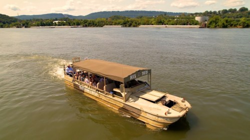 A boat with passengers navigates a river, surrounded by green hills and a clear blue sky.