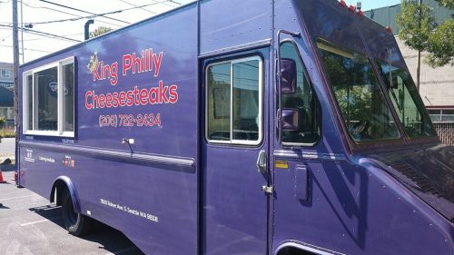 A purple food truck named "King Philly Cheesesteaks" parked in a lot, featuring a large window and menu details.