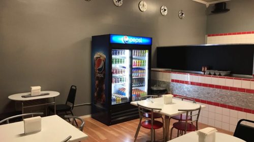 A small diner with tables, chairs, and a Pepsi vending machine against a gray wall.