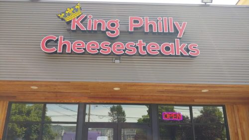 Sign for "King Philly Cheesesteaks" with a crown logo, featuring an "Open" neon sign in the window.