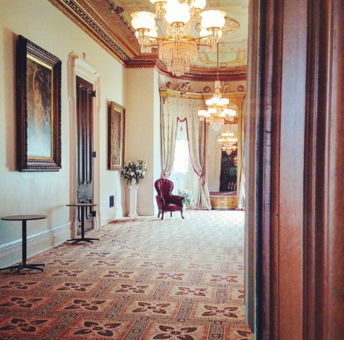 Elegant room with ornate chandeliers, a red armchair, and decorative paintings, featuring a patterned carpet.